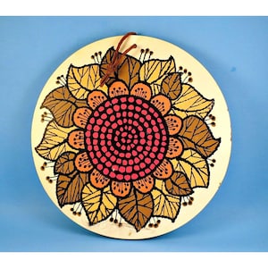 Może przedstawiać: Okrągła, kremowa dekoracja ścienna z motywem stylizowanego słonecznika. Słonecznik ma różowy środek z czarną spiralą, otoczony pomarańczowymi i brązowymi płatkami oraz żółtymi liśćmi. Brązowy skórzany sznurek do zawieszenia. Około 25 cm średnicy.