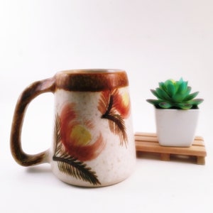 Può includere: Una tazza da caffè in ceramica con un bordo marrone e un corpo bianco. La tazza ha un motivo floreale dipinto su di essa in tonalità di arancione, giallo e marrone. La tazza è appoggiata su una superficie bianca.