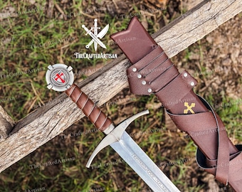 May include: A medieval sword with a silver blade and a brown leather-wrapped handle. The sword rests beside a brown leather sheath with a gold cross emblem. The sword's hilt features a shield with a red cross. TheCraftedArtisan logo is visible.