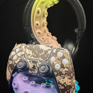 Könnte beinhalten: Ein Gaming-Controller mit Tarnmuster liegt auf einem lila-blauen, oktopusförmigen Ständer. Ein schwarzer Kopfhörer befindet sich über dem Controller. Die Oktopus-Tentakel sind gelb, rosa und lila.