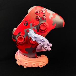 Könnte beinhalten: Ein rot-grau getarnter Videospiel-Controller mit einem hellvioletten und orangefarbenen Ständer im Oktopus-Design. Der Controller hat rote Knöpfe und einen schwarzen Hintergrund.