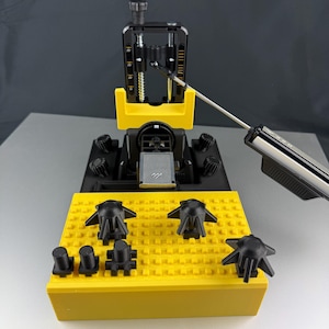 Könnte beinhalten: Ein gelb-schwarzes Präzisionswerkzeug mit einem silbernen Arm, der sich von oben erstreckt. Das Werkzeug hat eine Reihe schwarzer Knöpfe und eine gelbe Basis. Mehrere schwarze Komponenten sind auf der gelben Basis angeordnet.