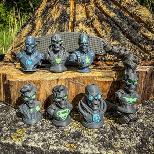 Könnte beinhalten: Eine Sammlung von sieben grauen, 3D-gedruckten Superhelden-Büsten im Totenkopf-Design mit leuchtend grünen und blauen Akzenten an den Brustemblemen und Augen. Die Figuren zeigen stilisierte Versionen von Iron Man, Captain America, Superman, Batman und dem Joker.