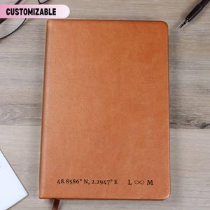 May include: A customizable, brown leather journal with the text "48.8586° N, 2.2947° E L ∞ M" printed at the bottom. The journal has a textured surface and a ribbon bookmark. The word "CUSTOMIZABLE" is in a pink banner at the top.