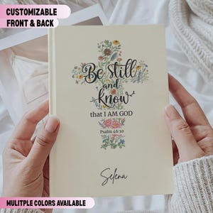 May include: Cream-colored journal with floral cross design and the text "Be still and know that I AM GOD" and "Psalm 46:10". The name "Selena" is written at the bottom. The journal is held by a person.