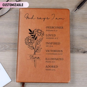 May include: A tan leather-like journal featuring a black floral illustration and the text God says I am, followed by a list of biblical affirmations including Overcomer, Loved, Inspired, Victorious, Illuminated, and Adored with scripture references.