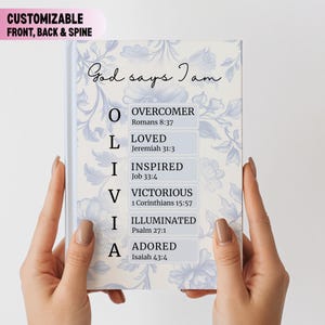 May include: A personalized journal with a floral design and the words "God says I am" in script. The journal's cover features the name "Olivia" spelled out with inspirational words and Bible verses. The journal is held by two hands.
