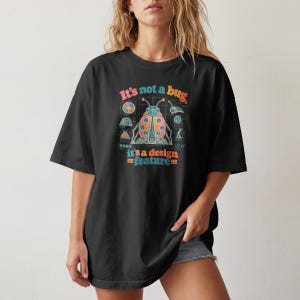 May include: A black short-sleeved t-shirt featuring a colourful graphic of a stylised ladybug with technical blueprint lines and diagrams. The text reads, It's not a bug, it's a design feature, in a multi-coloured, retro-inspired font style.