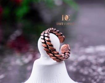 Può includere: Un anello color rame con un design intrecciato. L'anello è aperto nella parte inferiore e poggia su un piedistallo bianco. L'anello ha una superficie strutturata ed è realizzato in rame. Il testo "DMT Copper" è visibile sullo sfondo.