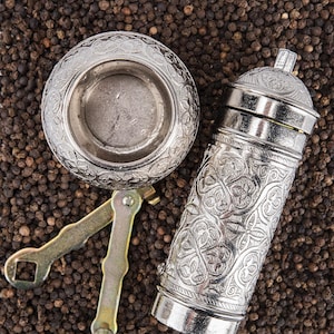 May include: A silver spice grinder and bowl with ornate floral patterns, alongside a small wrench, rest on a bed of black peppercorns. The grinder is cylindrical, and the bowl is round.