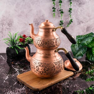 Könnte beinhalten: Eine traditionelle türkische Kupferteekanne mit aufwendigen floralen Mustern. Die Teekanne hat zwei Ebenen und Holzgriffe. Sie steht auf einem Holzbrett.