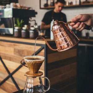 May include: A copper gooseneck kettle pouring hot water into a coffee pour-over setup. The setup includes a ceramic dripper and a glass carafe. The scene takes place in a coffee shop, with a barista in the background.