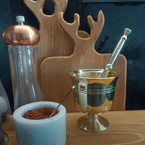 May include: A gold mortar and pestle set on a wooden surface. The mortar has a round base and a wide, shallow bowl. The pestle is long and slender, with a rounded end. The set is next to a small white container with red powder inside.