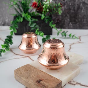 May include: Two copper salt and pepper shakers with a hammered finish, sitting on a wooden and marble chopping board.