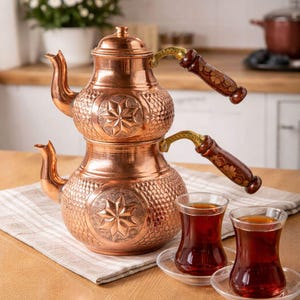 May include: A copper tea set with a two-tiered teapot and two glasses of tea. The teapot has a starburst design and wooden handles. The tea glasses are clear and sit on small saucers. The set is on a wooden table.