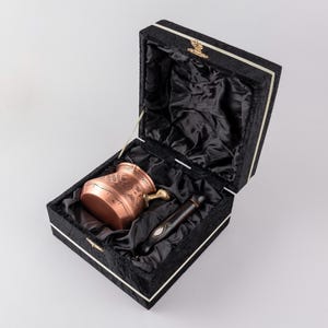Op de afbeelding: Een koperen koffiepot met ingewikkelde gravures wordt getoond in een met zwart fluweel gevoerde doos met een houten handvat.