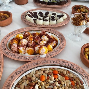 May include: A table setting with a variety of Turkish dishes, including a plate of small, white, round desserts topped with chocolate, a plate of small, round, orange-colored desserts, a plate of mixed nuts, and a glass of yogurt with fruit.