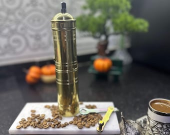 Pode incluir: Um moedor de café manual dourado com uma alça preta, sentado em uma bandeja de mármore branco com grãos de café. Uma xícara de café e um pequeno recipiente de prata também estão presentes, sugerindo um tema de preparação de café.
