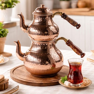 May include: A copper tea set with a two-tiered teapot and a glass of tea. The teapot has a hammered texture and wooden handles with floral designs. The tea is served with sugar cubes and mint on a small plate.