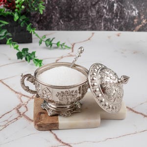Könnte beinhalten: Eine silberne Zuckerdose mit aufwendigen floralen Mustern, gefüllt mit weißem Zucker und einem kleinen Löffel. Der Deckel ist teilweise geöffnet. Die Dose steht auf einem Servierbrett aus Marmor und Holz.