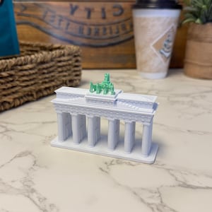 May include: A white miniature replica of the Brandenburg Gate, a neoclassical monument. The model features detailed columns and a green Quadriga sculpture on top. The miniature is a detailed architectural model.