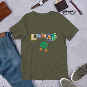 Könnte beinhalten: Ein grünes T-Shirt mit kurzen Ärmeln mit dem Text "Lucky AF" in bunten Buchstaben und einem Cartoon-Kleeblatt mit einem lächelnden Gesicht.