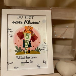 Könnte beinhalten: Ein gerahmter Druck mit dem Text "DU BIST erste Klasse!" und einer Illustration eines Kindes, das ein "5"-Schild hält. Der Druck enthält zusätzlichen Text und ist von mehreren Personen signiert. Ein herzförmiges Objekt liegt darunter.