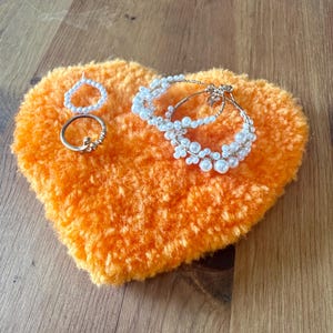 以下が含まれることがあります： オレンジ色のハート型のふわふわした装飾品で、いくつかのジュエリーが付いています。ジュエリーには、花の模様のリング、ビーズリング、パールのフープイヤリングが含まれます。ジュエリーは金と白です。