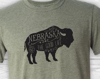 May include: A green t-shirt with a black graphic of a buffalo silhouette. The graphic includes the text "Nebraska, Since 1867, The Good Life".
