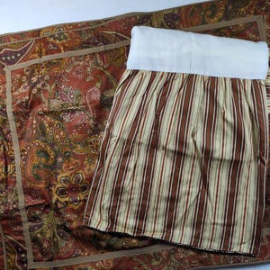 May include: A bed skirt featuring a white top panel and a pleated drop with vertical stripes in shades of brown, cream, and tan. It is layered over a flat fabric piece decorated with a complex, multi-colored paisley pattern in rust, green, and gold.