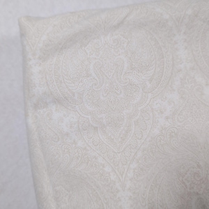 May include: A folded textile fabric featuring a light beige and white damask pattern. The design consists of intricate, symmetrical floral and scrollwork motifs repeated across the surface, creating a soft, neutral, and detailed visual texture.