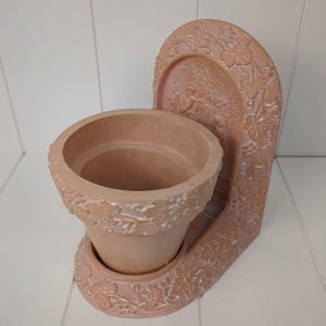 May include: A terracotta-colored planter with a decorative arched back. The planter has a round pot with leaf and vine detailing. The arched back features a relief of a figure and more leaf and vine designs.