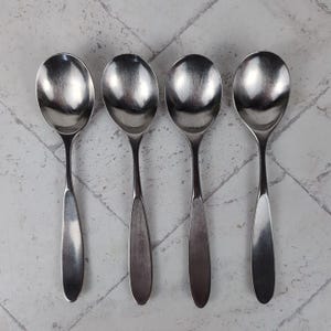 May include: Four stainless steel soup spoons arranged in a row. Each spoon has a rounded bowl and a long, slender handle. The spoons are made of a shiny, silver-colored metal and are set against a light gray and white background.