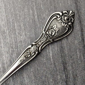 May include: A silver spoon with an ornate, floral design on the handle. The handle is decorated with raised roses and leaves.