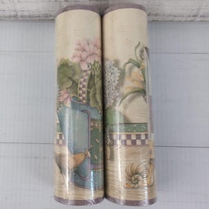 May include: Two rolls of wallpaper border featuring a cream background with a repeating pattern of blue watering cans, pink flowers, green foliage, and a small snail. The design includes a checkered border detail and a light tan wood-grain texture.