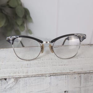 May include: Vintage eyeglasses with a black upper frame and silver-colored lower frame. The temples are black with decorative silver accents. The lenses are clear, and the nose pads are beige. The glasses are set against a white, textured surface.