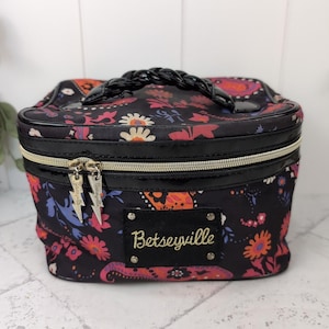 Puede incluir: Bolsa de cosméticos negra con un estampado floral en tonos rosa, naranja y azul. La bolsa tiene un asa trenzada negra, una cremallera dorada y la palabra "Betseyville" en escritura dorada en la parte delantera.