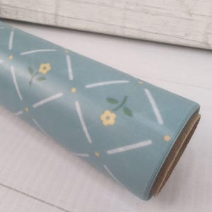 May include: A roll of light blue contact paper featuring a repeating pattern of white diagonal lines forming a diamond grid, with small yellow flowers and tiny yellow dots centered within each diamond shape across the smooth surface.