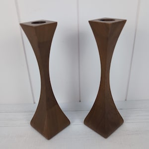 May include: A pair of tall, dark brown wooden candle holders featuring a smooth, hourglass-shaped silhouette with a square base and a square top. Each holder has a circular opening at the center of the top surface for holding a single candle.