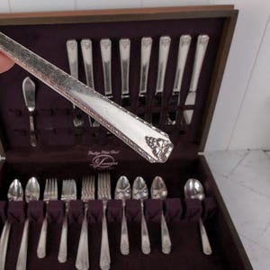 May include: A silver-colored flatware set with ornate floral detailing, displayed in a wooden case with a dark purple interior. The set includes knives, forks, spoons, and a butter knife. The flatware is arranged neatly in the case.