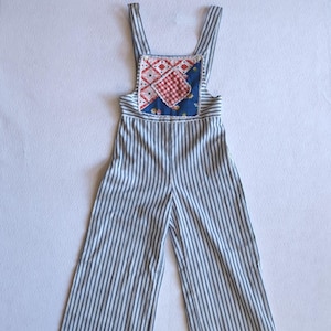 May include: Vintage-style striped overalls with a patchwork bib. The overalls feature vertical blue and white stripes on the legs and a bib with a red and white checkered pattern, floral fabric, and a blue background. The straps are white.