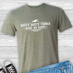 Puede incluir: Una camiseta verde con un gr&aacute;fico de p&aacute;jaro blanco y el texto "Rusty, Dusty Things Make Me Happy".