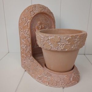 May include: A terracotta-colored planter with a decorative arch and pot. The arch features a relief of a figure and grape vine designs. The pot has a wide rim with a grape vine pattern. The planter is designed to hold a small plant.