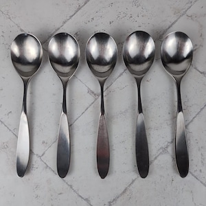 May include: Five stainless steel soup spoons arranged in a row. Each spoon has a rounded bowl and a long, slender handle. The spoons are reflective, showing the light and the surrounding environment. The spoons are on a light gray and white patterned surface.