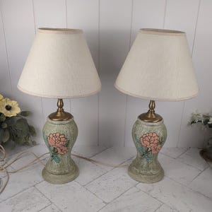 May include: Pair of vintage table lamps with off-white fabric shades. The lamps have a green base with a floral design in pink and blue. The base has a brass-colored neck and a cream-colored shade.