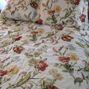 May include: A floral bedding set featuring a white comforter and two matching pillowcases. The pattern includes red, yellow, and blue flowers with green leaves. The bedding is on a wooden bed frame.