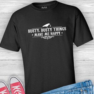 Puede incluir: Camiseta negra con texto blanco que dice "Rusty, Dusty Things *Make Me Happy*" y una peque&ntilde;a silueta blanca de un p&aacute;jaro sobre el texto.