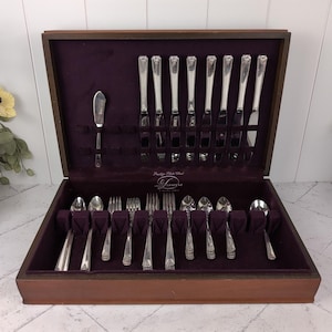 May include: A vintage silverware set in a wooden case with a deep purple velvet lining. The set includes knives, forks, spoons, and a butter knife, all with ornate handles. The case is open, displaying the arranged cutlery.