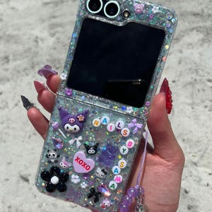 Könnte beinhalten: Eine transparente, glitzernde Handyhülle im Klappdesign, verziert mit verschiedenen Charms, darunter ein lila Kuromi-Charakter, eine schwarze Blume und das Wort "XOXO" auf einem Herz. Die Hülle hat auch einen Perlenriemen.