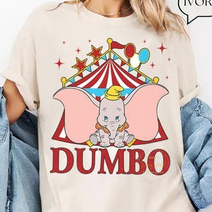 May include: Ivory t-shirt with a Dumbo graphic. The design features a circus tent, stars, and balloons. The word "DUMBO" is printed in red, with a cartoon elephant in the center. This casual t-shirt is suitable for everyday wear.